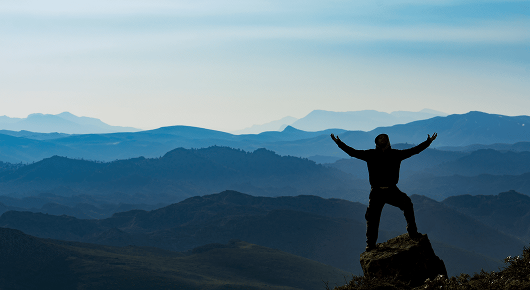 Photo: sillhouette of a man standing on a mountain lookout with his hands held up in thankfulness.