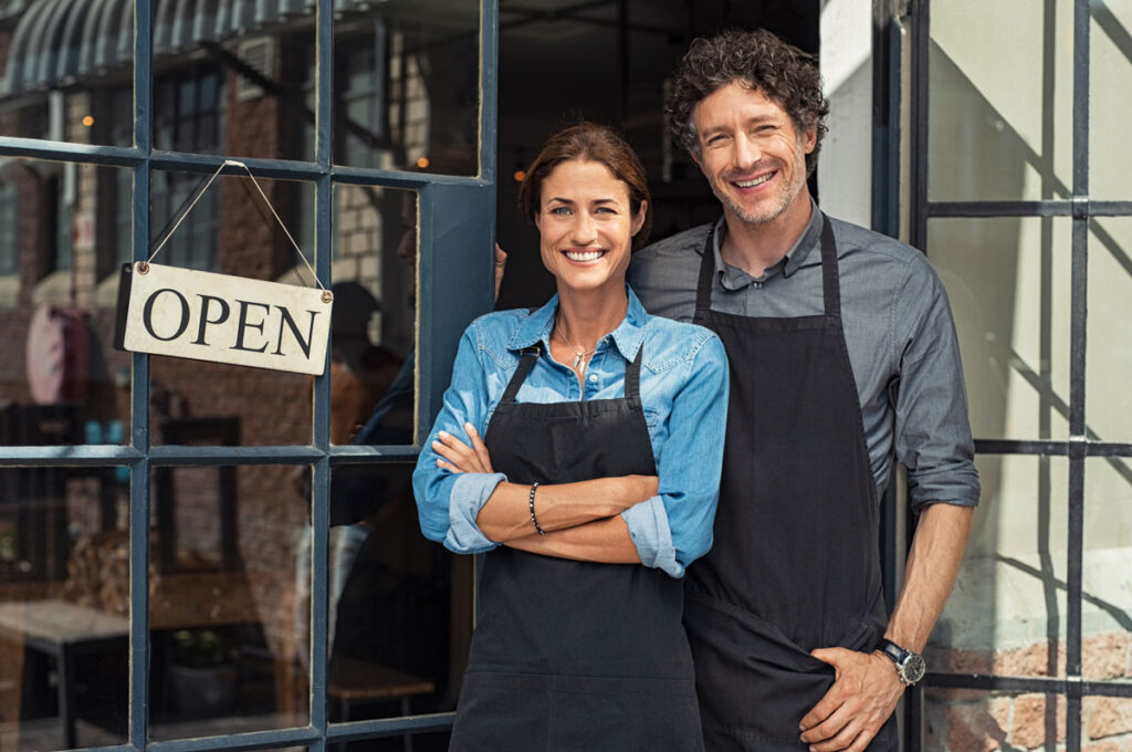 Two small business owners smiling and looking at camera while standing at entrance door.