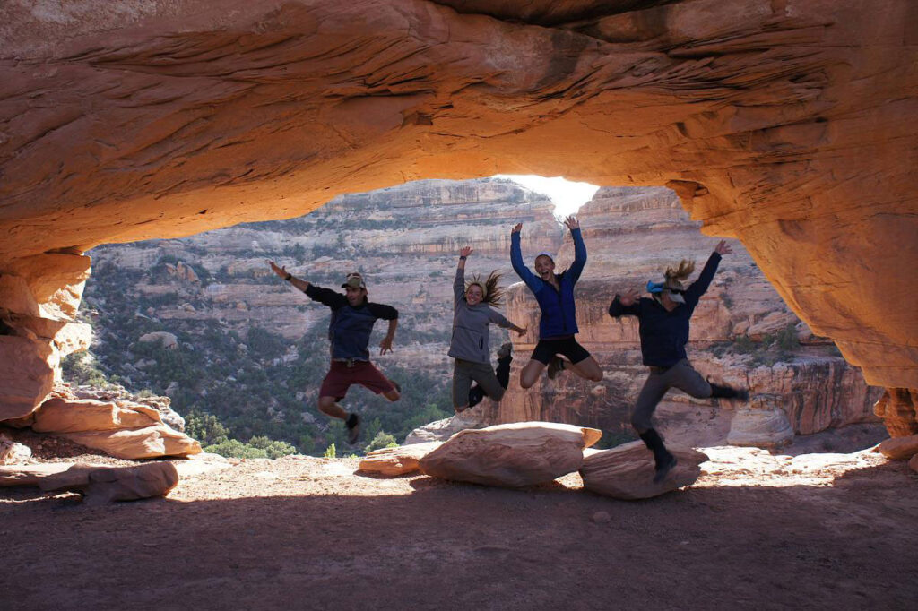 CMC Outdoor Recreation Leadership students jump under a sandstone arch in the desert.